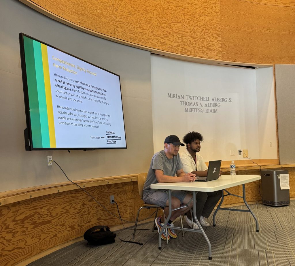 A light-skinned person in a baseball cap and a dark-skinned person sit at a table in front of a presentation screen in a meeting room at Ballard Library. 